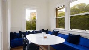 Una habitación con una mesa redonda blanca, asientos tapizados en azul y amplios ventanales con vistas a zonas verdes y edificios.