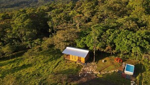 Une petite cabane jaune, surmontée d’un toit en métal, entourée d’une végétation luxuriante et avec une montagne en arrière-plan.