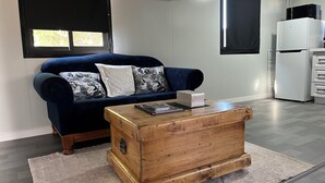Una sala de estar con un sofá azul, un cofre de madera, un refrigerador blanco y una ventana con persianas.