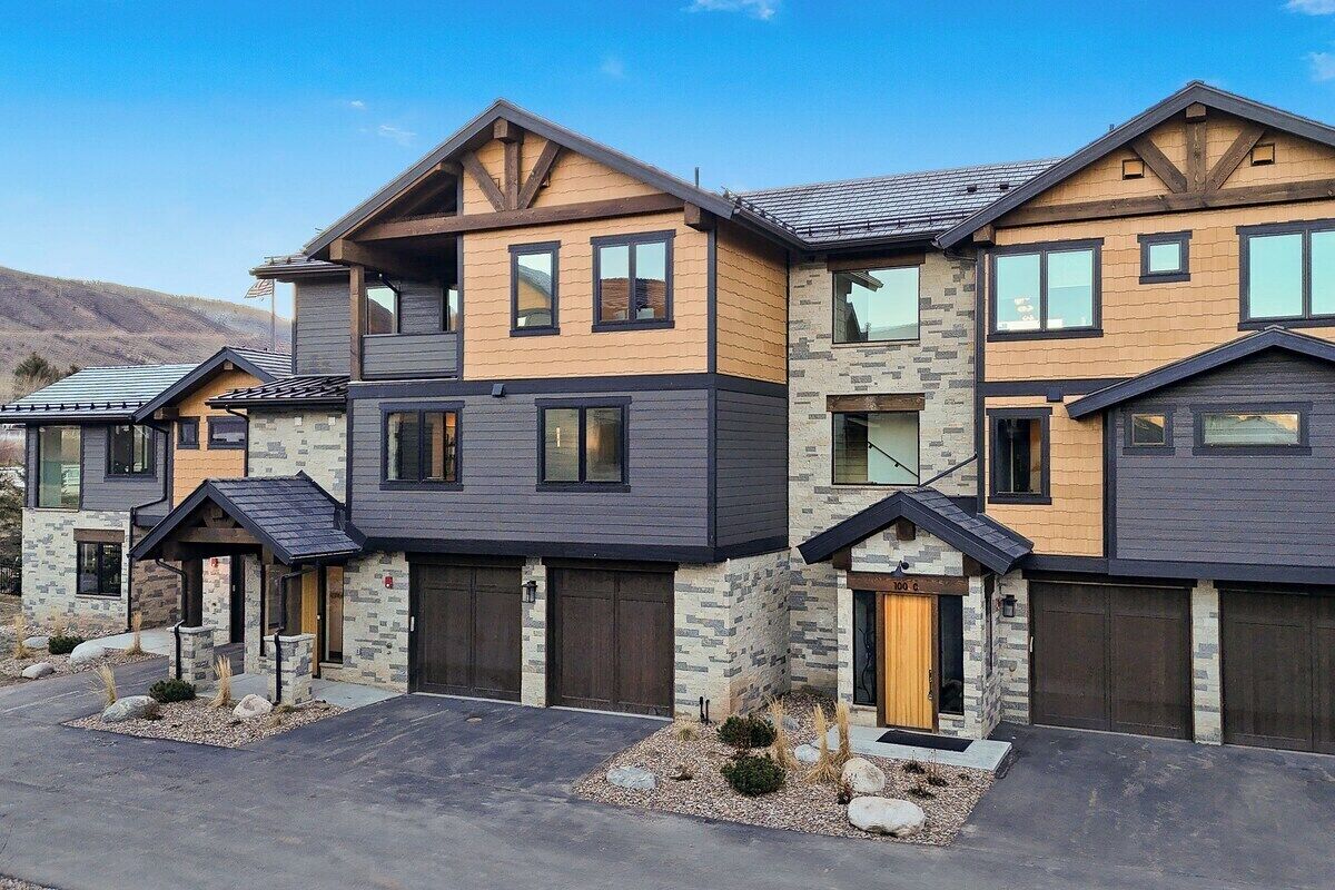 Une rangée de maisons modernes à plusieurs étages, aux façades en pierre et en bois, avec des portes de garage, sous un ciel dégagé.