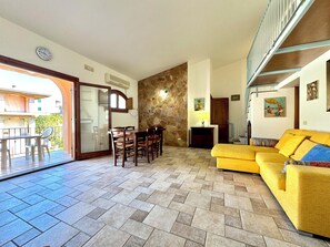 Una sala de estar con un sofá amarillo, una mesa de comedor con sillas, un muro de piedra y una escalera.