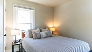 Un dormitorio ordenado con una cama, dos mesitas de noche, una lámpara y una ventana.