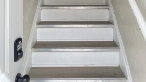 Una escalera con escalones blancos y barandilla, que lleva a una puerta con mirilla y timbre.
