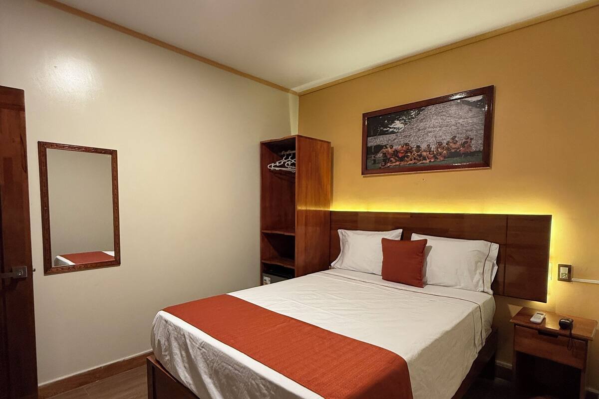 Une chambre d’hôtel avec un lit, une armoire en bois, une table de chevet, une commode, un miroir et un tableau accroché au mur.
