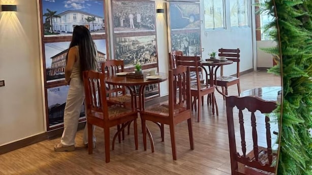 A dining area with wooden chairs and tables, a person standing near a wall with framed pictures, and a large window with a view. - Hotel Yagua Iquitos (Belen)