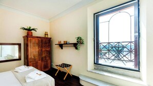 Een hotelkamer met een bed, een houten garderobe, een kleintje tafeltje met een plant, een raam met uitzicht op een balkon en een plank met decoratieve objecten.