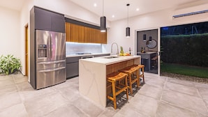 A modern kitchen with a stainless steel refrigerator, wooden cabinets, a central island with stools, pendant lighting, and a view of a garden through a glass door.
