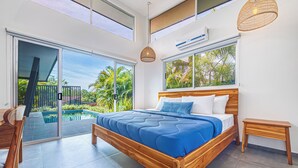 A modern bedroom with a wooden bed, a glass door leading to a pool, and a view of palm trees.