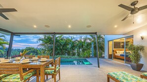 A spacious outdoor dining area with a pool, wooden furniture, and a view of the sky and greenery.