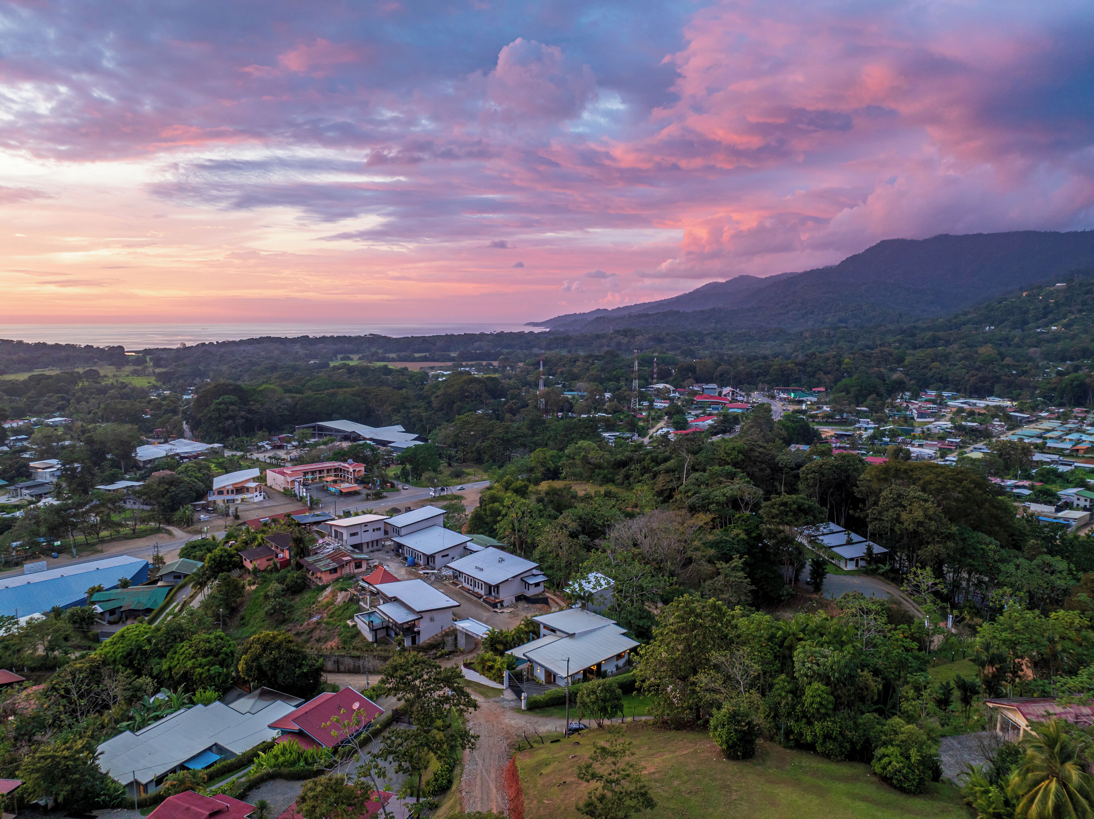 An aerial view of a coastal town with a mix of residential and commercial buildings, surrounded by lush greenery and a mountainous backdrop.