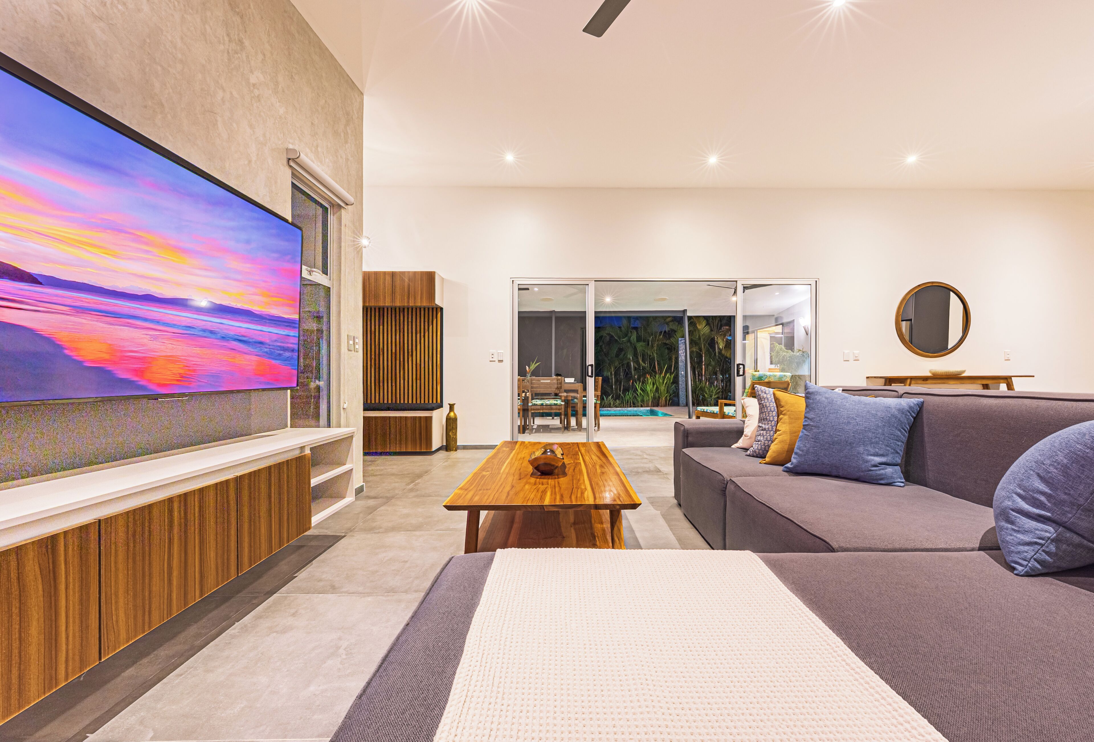 Una sala de estar moderna con un televisor de pantalla plana grande, una mesa de centro de madera, un sofá gris y vistas a una piscina a través de puertas de vidrio.