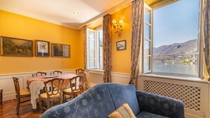 A room with a dining table, chairs, a sofa, and a large window with a view of a lake and mountains. - Casa Orta apartment with lake view (Orta San Giulio)