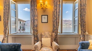 A room with a patterned armchair, two sofas, a wall-mounted light fixture, and large windows with curtains. - Casa Orta apartment with lake view (Orta San Giulio)