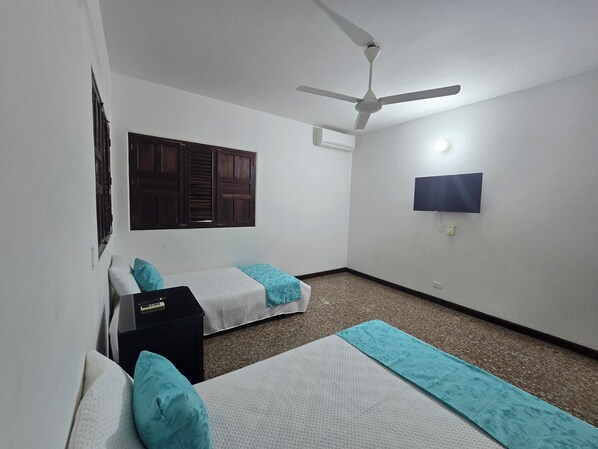 Una habitación con dos camas, un ventilador de techo, un televisor montado en la pared y una ventana con contraventanas de madera.