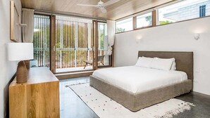 Een moderne slaapkamer met een groot bed, een houten nachtkastje, een plafondventilator en uitzicht op de natuur door een schuifdeur.