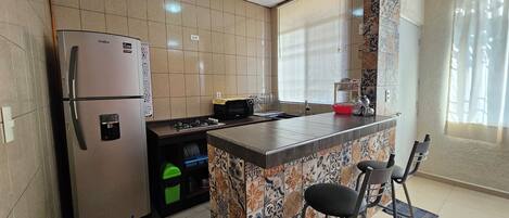Una cocina con refrigerador, estufa y microondas. Hay una ventana con persianas y una silla cerca del fregadero.