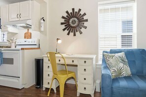 A compact kitchen with white cabinets, a stove, and a yellow chair. There's a blue couch with a leaf-patterned pillow and a wall clock. - Uptown Cottage l Walk to DNTN l Dogs OK (St. Augustine)