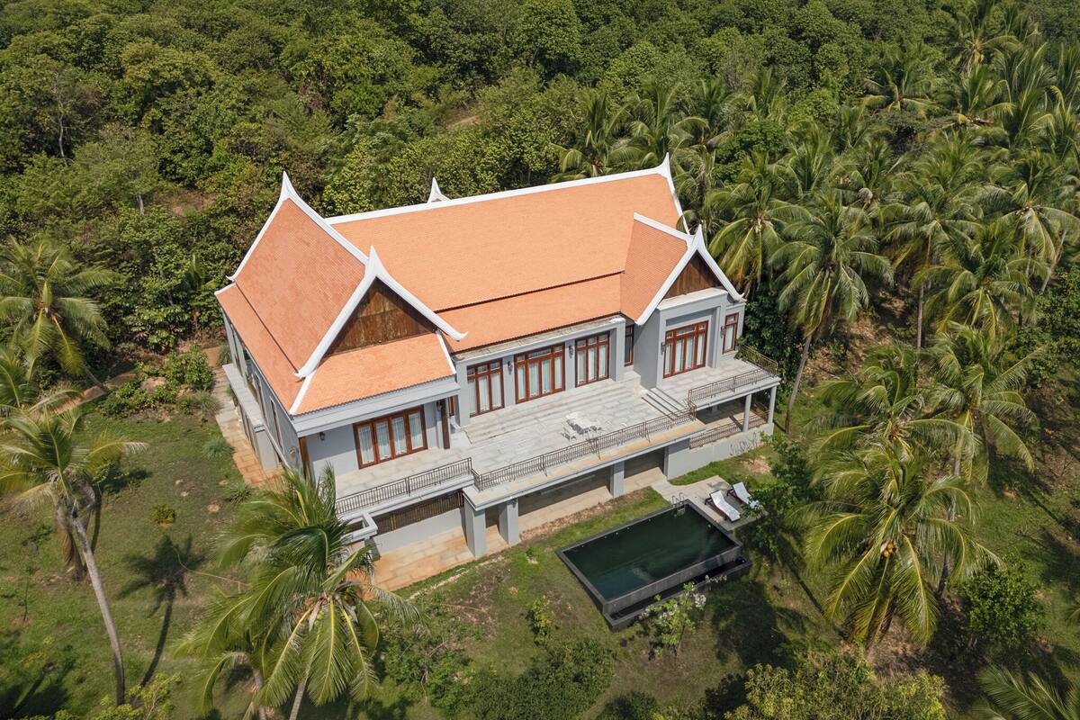 A large, two-story house with a tiled roof, surrounded by lush greenery and palm trees.