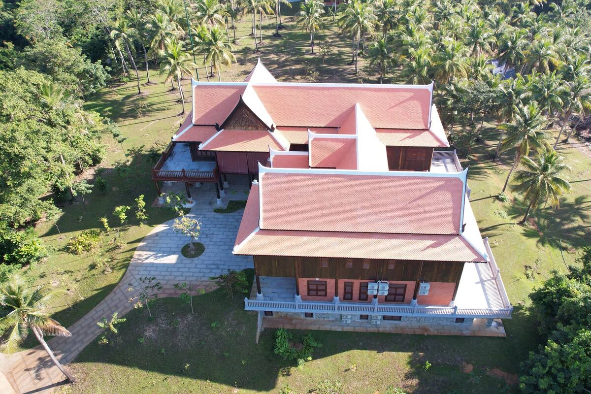 A large, multi-story building with a red roof, surrounded by lush greenery and palm trees.