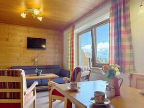 A cozy room with a wooden ceiling, a television mounted on the wall, a large window with colorful curtains, a sofa, a table with cups and a pitcher, and a view of a mountain landscape. - Katschberg 26 by Interhome (Sankt Michael im Lungau)