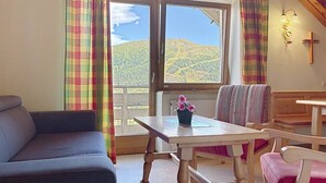 A room with a sofa, a table with a flower vase, a bench, and a view of a mountain landscape through a window. - Katschberg 26 by Interhome (Sankt Michael im Lungau)