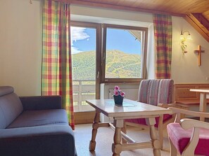 A room with a sofa, a table with a flower vase, a bench, and a view of a mountain landscape through a window. - Katschberg 26 by Interhome (Sankt Michael im Lungau)