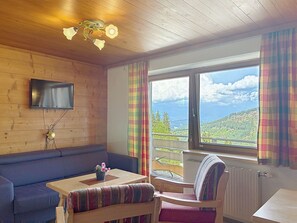 A cozy room with a wooden ceiling, a flat-screen TV mounted on the wall, a sofa, a table with a chair, and a window with a view of a mountainous landscape. - Katschberg 26 by Interhome (Sankt Michael im Lungau)