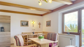 Um quarto acolhedor com uma mesa de jantar de madeira, cadeiras, uma cama com lençóis brancos, uma janela com vista para árvores e uma cruz na parede.