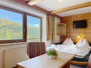 A hotel room with a bed, a table with a plant, a chair, a television mounted on the wall, and a window with a mountain view. - Katschberg 23 by Interhome (Sankt Michael im Lungau)