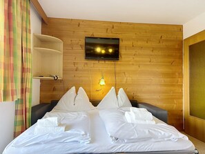 A hotel room with a wooden wall, a flat-screen TV mounted above a bed, a bedside table, and a window with curtains. - Katschberg 23 by Interhome (Sankt Michael im Lungau)