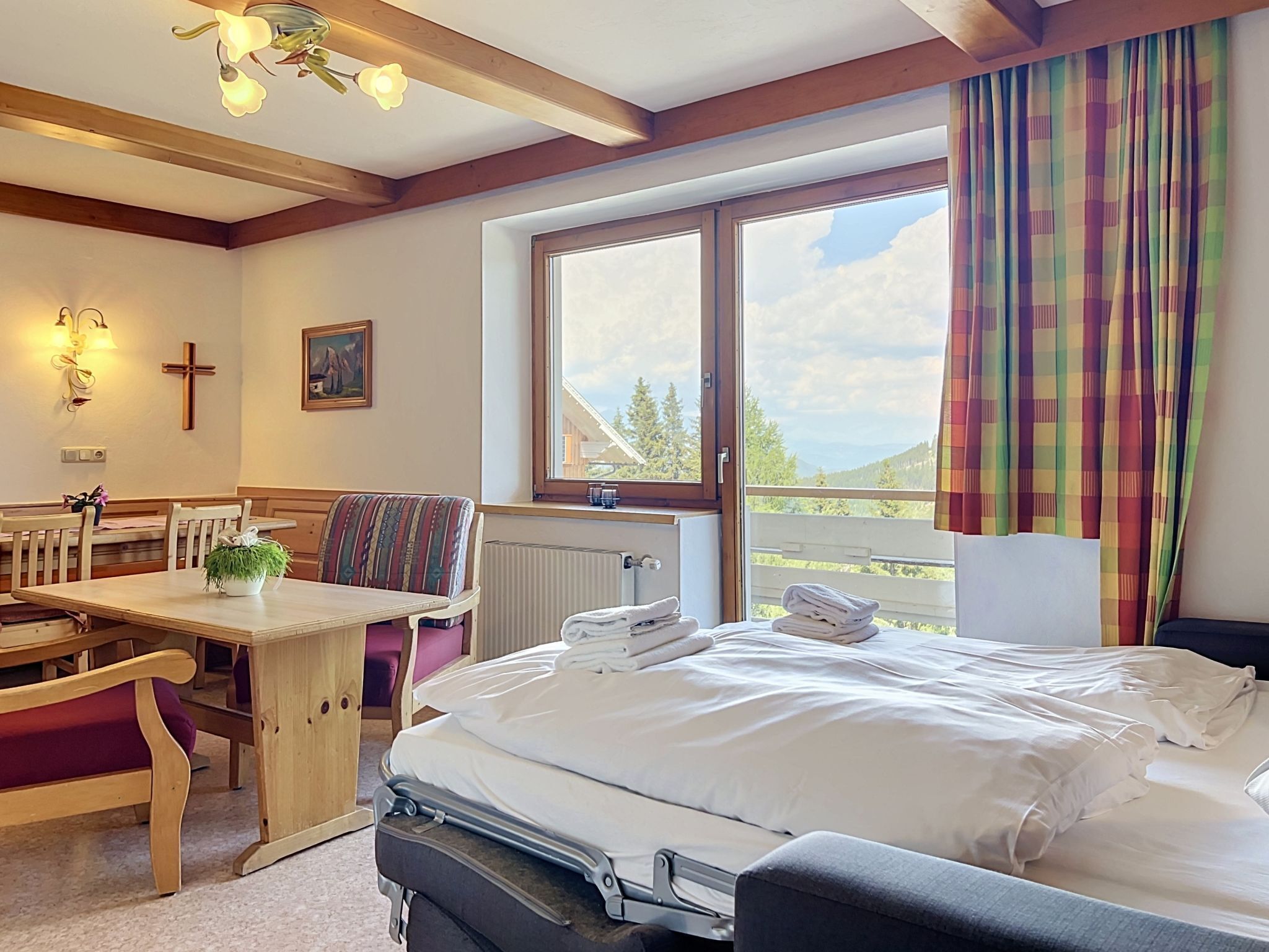 A hotel room with a bed, a table with chairs, a window with curtains, a radiator, and a cross on the wall.