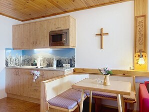 Une petite cuisine équipée d’un four à micro-ondes, d’un évier et d’une cuisinière. Un crucifix en bois est accroché au mur, et une petite table avec un banc se trouve dans la pièce.