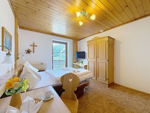 A hotel room with a wooden ceiling, a bed with white linens, a television, a wardrobe, and a window with a view of trees.