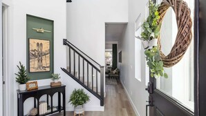 Un pasillo con una escalera, un cuadro en la pared, una consola con plantas y una corona de mimbre en la puerta.