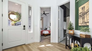 Un pasillo con suelo de madera, una puerta adornada con un arreglo floral, un ventilador de techo, una cama con una colcha roja y una pared con un cuadro enmarcado.