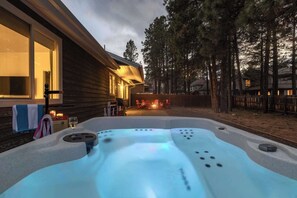 Un bain à remous dans un jardin, avec une maison en bois et un foyer en pierre en arrière-plan.
