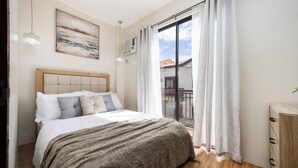 Une chambre à coucher avec un lit, une table de chevet, une fenêtre avec des rideaux, un climatiseur fixé au mur et un tableau au-dessus du lit.