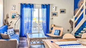 Ett vardagsrum med utsikt över havet, en soffa med blå kuddar, ett soffbord i trä och en trappa med blå detaljer.