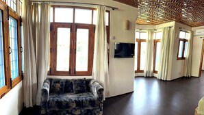 Uma sala de estar ampla, com piso de madeira escura, um sofá com almofadas estampadas e grandes janelas com cortinas.