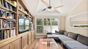 Ein Wohnzimmer mit einer Holzbücherreolen, einem Flachbildfernseher, einem Deckenventilator, einem Sofa und einem Fenster mit Jalousien.