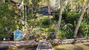 Eine Holzbrücke über einen Teich, die zu einem Haus inmitten üppiger Vegetation und Bäume führt.