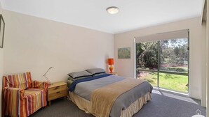 A bedroom with a bed, a nightstand, a lamp, a chair, and a large window with a view of greenery. - The Ash, Sandy Point (Sandy Point)