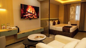 A hotel room with a bed, a TV displaying a fireplace, a dining area with a table and chairs, and a small kitchenette. - Emboita Hotels ltd (Nakuru)
