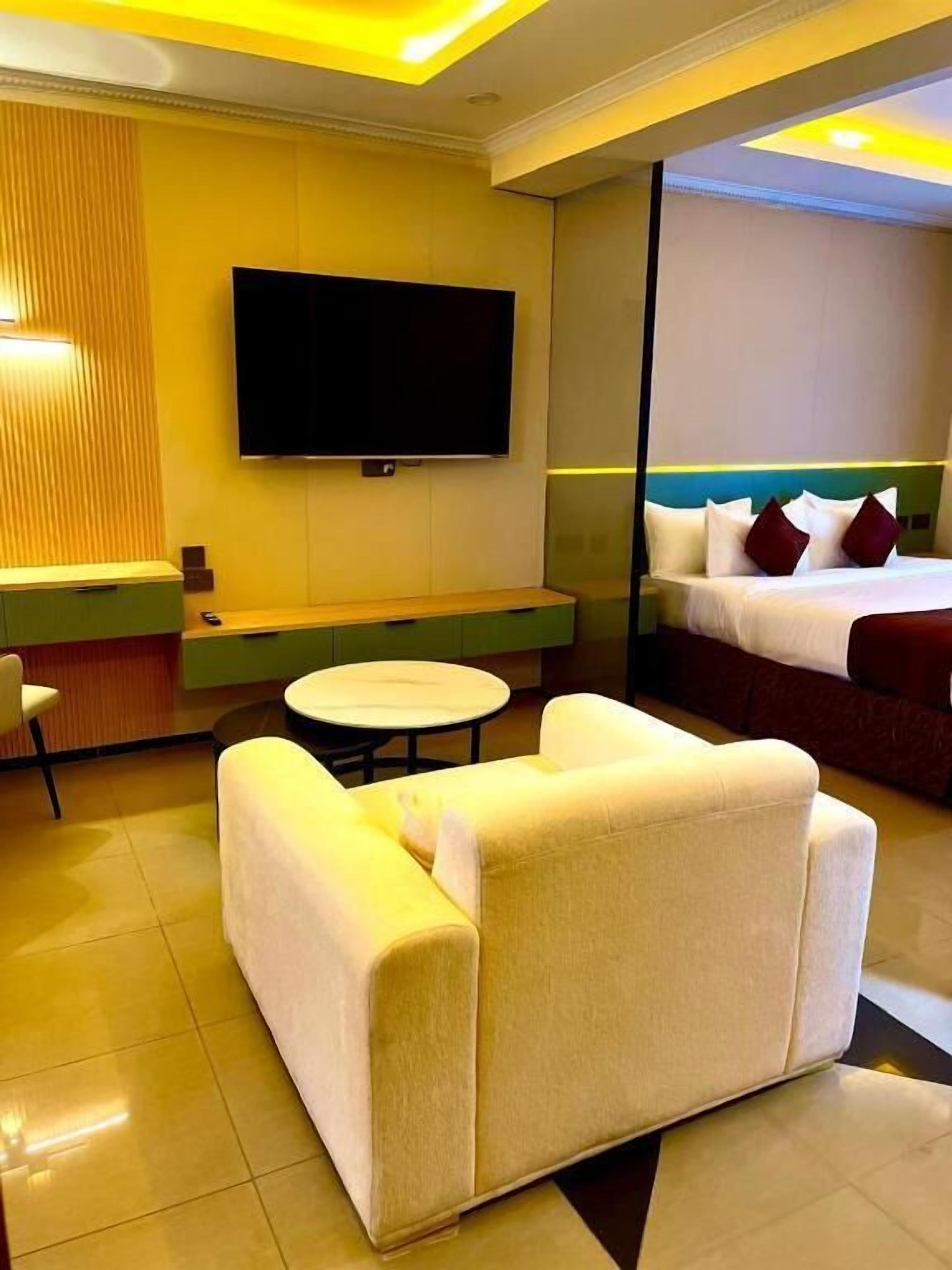 A hotel room with a flat-screen TV mounted on the wall, a bed with white bedding and red pillows, a sofa, a small round table, and a green console table.