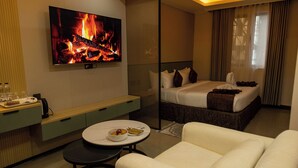 A hotel room with a bed, a TV displaying a fire, a dining table with a bowl of fruit, and a sofa. - Emboita Hotels ltd (Nakuru)