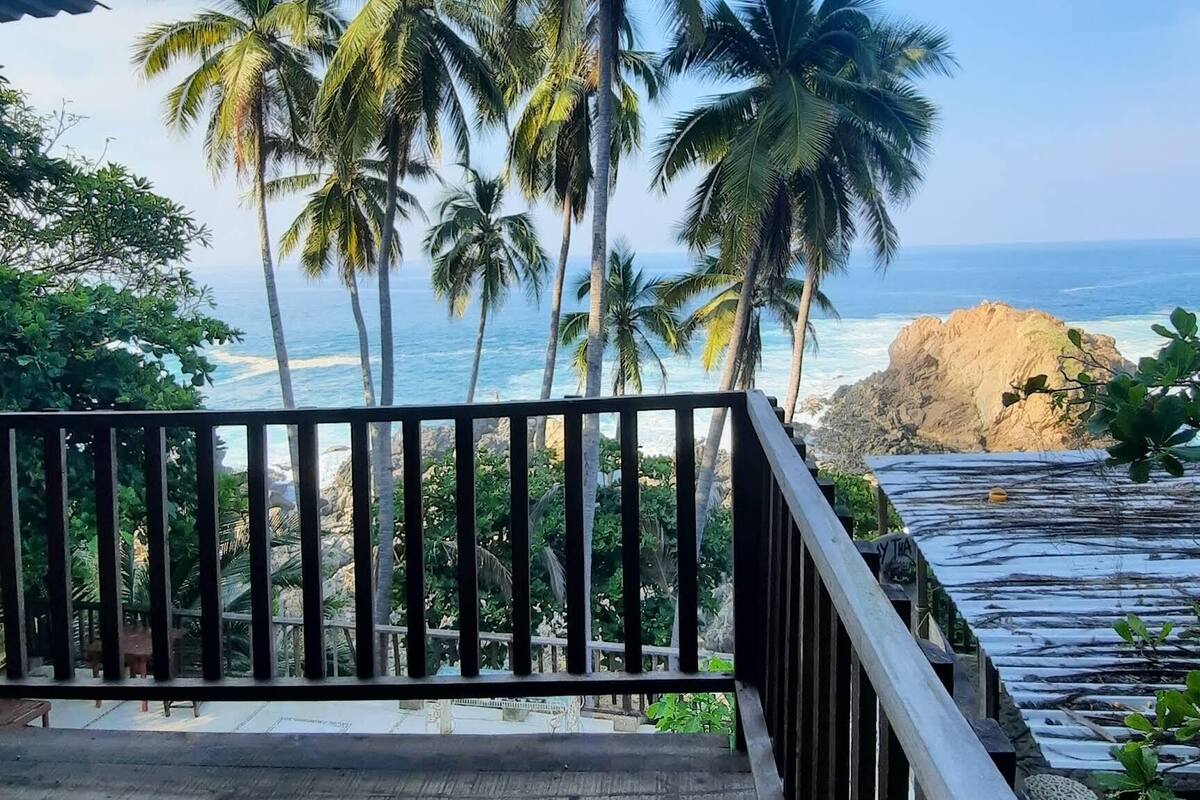 A wooden deck with a railing, overlooking a beach with palm trees and a rocky outcrop.