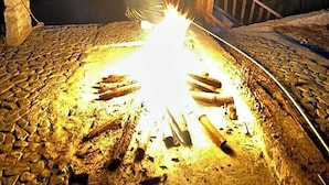 A campfire with logs arranged in a circle, surrounded by a stone-paved area with a wooden bench and a stone wall. - Cocos camp (Acapulco)