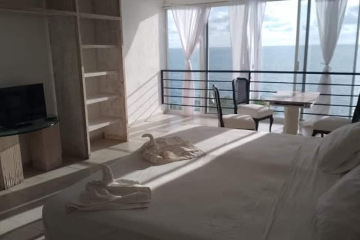 A hotel room with a bed, a TV, a balcony with a table and chairs, and a view of the sea.