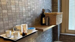 Une salle de bain moderne avec un mur recouvert de carreaux de pierre, une étagère en bois sur laquelle est posé un panier, et deux tabourets ronds.