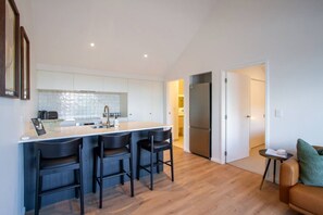 A modern kitchen with a central island, bar stools, a refrigerator, and a dining area with a sofa and a small table. - Modern Central City Apartment | King Bed (Christchurch)